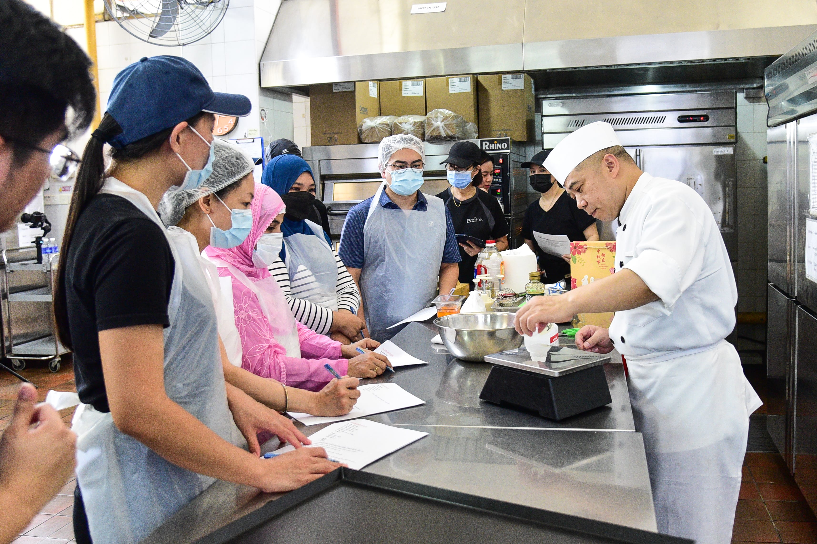 A group of individuals gathered around a kitchen island, watching a baker demonstrate baking techniques.